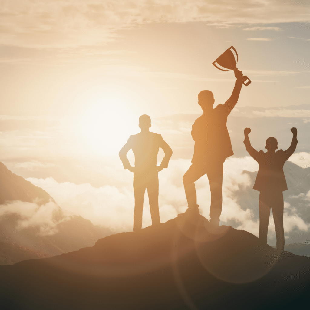 Three people on a mountain peak at sunset creating a silhouette. One person is holding a trophy overhead in celebration of their achievement while another has both hands raised above their head in celebration.